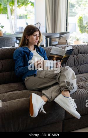 Frau im Alter von 20 Jahren, die zu Hause auf dem Sofa sitzt, eine Jeansjacke trägt, einen Becher hält und Taschenbuch liest Stockfoto