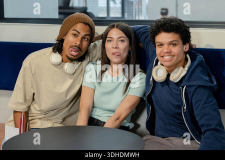 Afroamerikanische Freunde sitzen auf blauer Bank in der Lounge mit weißen Kopfhörern, Beanie und Notebook Stockfoto