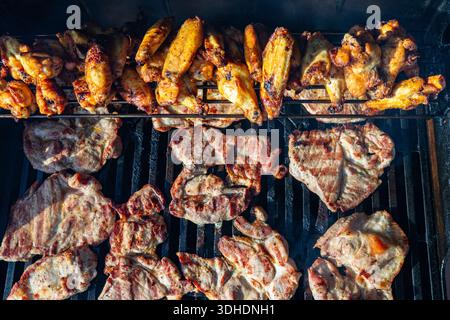 Lebendiger Grill im Freien mit brausenden Hühnerflügeln auf einem Gestell darüber und saftigen Schweinerippchen auf dem Grill, die rauchige Aromen, kräftige Aromen und ein einfangen Stockfoto