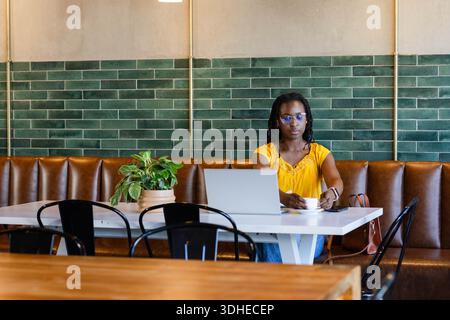 Frau mit gelber Bluse und Brille, die am Kaffeetisch sitzt, mit silbernem Laptop mit Tasse Stockfoto
