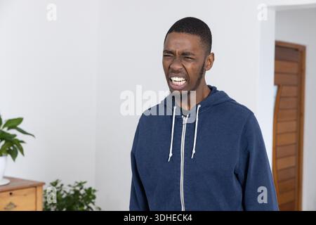 Erwachsener Afroamerikaner grimmend mit geschlossenen Augen, im Zimmer in Navy Hoodie in der Nähe der Kommode Stockfoto