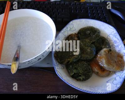 Eine Schüssel Congee mit Essstäbchen neben einem Teller mit traditionellem chinesischen grünen Reiskuchen und gebratenem Gebäck Stockfoto