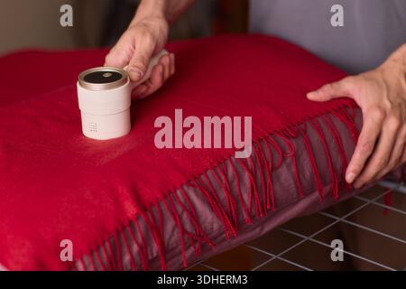 Hände reinigen roter Wollschal mit Fusselrolle über Fransenstoff, Nahaufnahme Textur, warme Schlafzimmerbeleuchtung, vorsichtige Bewegung, Entfernung von Tierhaaren Stockfoto