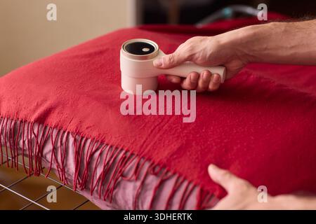 Hände reinigen roter Wollschal mit Fusselrolle über Fransenstoff, Nahaufnahme Textur, warme Schlafzimmerbeleuchtung, vorsichtige Bewegung, Entfernung von Tierhaaren Stockfoto
