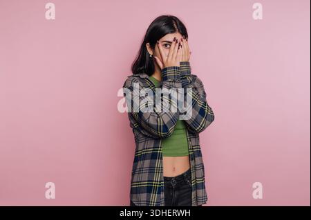 Junge Frau, die das Gesicht mit Händen vor rosa Hintergrund bedeckt. Stockfoto