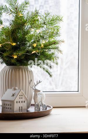Gemütliches Winterstillleben mit Tannenzweigen in einer weißen Vase, Keramikhaus und Hirschfigur auf einer Fensterbank mit weichem Tageslicht und festlichem atmos Stockfoto