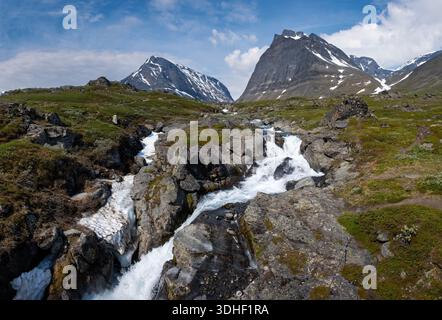 Dynamischer Blick auf einen kalten Bergfluss, der auf dem Kungsleden Trail über Felsen stürzt, mit dem dunklen, pyramidenförmigen Massiv der Kebnekaise im Hintergrund Stockfoto