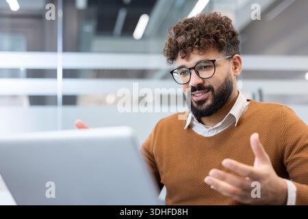 Junger bärtiger Mann mit lockigen Haaren und Brille, der auf einem Laptop ein Online-Business-Video anruft, aktiv kommuniziert und Gesten von einem modernen Büroarbeitsplatz aus bewegt Stockfoto