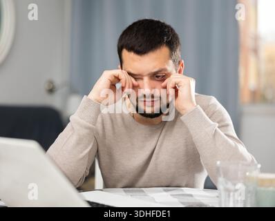 Junger Mann, der müde Augen reibt, während er zu Hause am Computer arbeitet Stockfoto