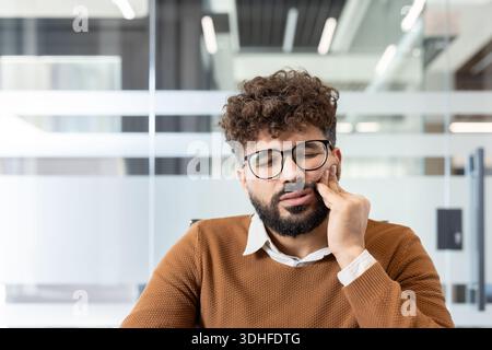 Mann mit Brille und lockigem Haar schließt Augen in Unbehagen, hält seine schmerzende Wange, während er Zahnschmerzen und Mundgesundheit Probleme in einer modernen Büroumgebung erlebt Stockfoto