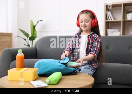 Mädchen mit Kopfhörern, die ihren Rucksack für die Schule auf dem Sofa zu Hause packen. Leerzeichen für Text Stockfoto