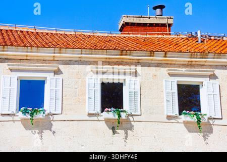 Ein weißes Steingebäude mit gekacheltem Dach steht vor klarem Himmel. Drei Fenster mit weißen Fensterläden und Blumenkästen verleihen mediterranen Charme Stockfoto