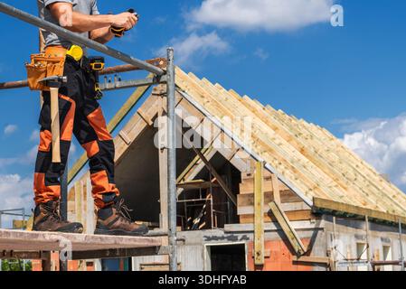 Ein Arbeiter steht auf einem Gerüst und bereitet sich darauf vor, das Dach unter klarem Himmel zu installieren. Die Werkzeuge sind am Gürtel des Werkers sichtbar. Stockfoto