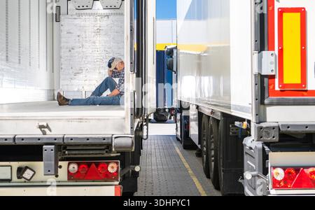 Müder Trcuker sitzt tagsüber auf dem Boden eines Lkw-Anhängers, während er in der Nähe anderer Lkw in einem Laderaum parkt. Stockfoto