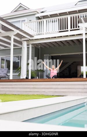 Frau, die auf Deck am Pool sitzt und Arme in rosa, dunkle Leggings mit Tanktop streckt Stockfoto