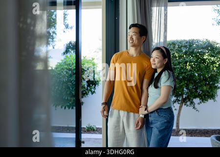 Asiatisches Paar, das an der Glasschiebetür steht, Hände hält, Frau mit rosafarbenem Stirnband, Kopierraum Stockfoto