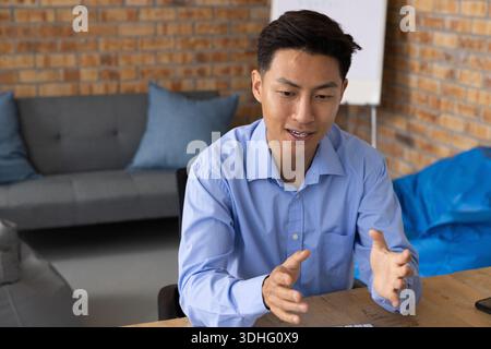 Asiatischer Mann in hellblauem Hemd, der beim Erklären am Tisch in der Bürolounge mit Keyboard Stockfoto