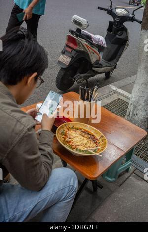 Rückansicht eines Mannes, der sitzt und isst, während er auf sein Handy schaut. Chengdu. China Stockfoto