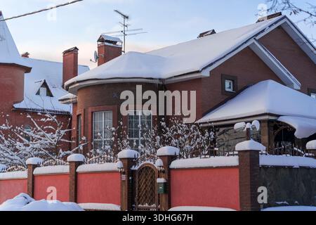 Zweistöckiges Privathaus rotem Backstein mit schneebedecktem Dach und Zaun im Winterlicht. Gemütliche Vorstadtarchitektur unter frischem Schnee, aufgenommen mit Sony A7 IV Stockfoto