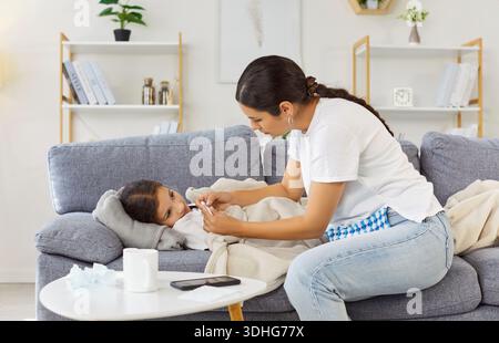 Die Mutter misst die Temperatur des kranken Kindes auf dem Sofa, pflegt und tröstet Stockfoto