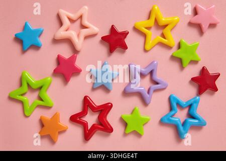 Eine farbenfrohe, flache Schicht aus glänzenden, sternförmigen Dekorationen auf einem sanften rosa Hintergrund, die eine verspielte, fröhliche Atmosphäre schaffen. Weihnachtsgrußkarten Stockfoto