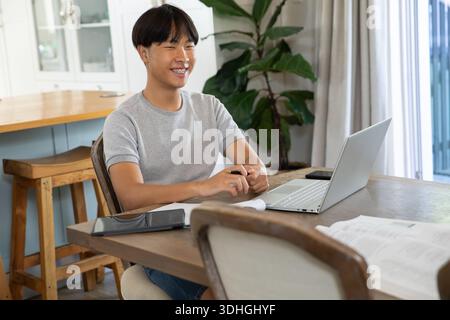 Asiatischer Mann, der zu Hause am hölzernen Esstisch sitzt, Laptop und Tablet benutzt, Stift hält Stockfoto