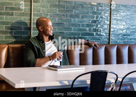 Mann lehnt sich zurück in einer braunen Lederkabine im Café und hält einen Take-out-Becher mit geschlossenem Laptop gestapeltem Telefon Stockfoto