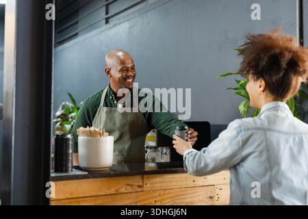 Afroamerikanischer Barista mit Schürze überreicht der Frau in einer Jeansjacke am Holztheken eine Tasse zum Mitnehmen Stockfoto