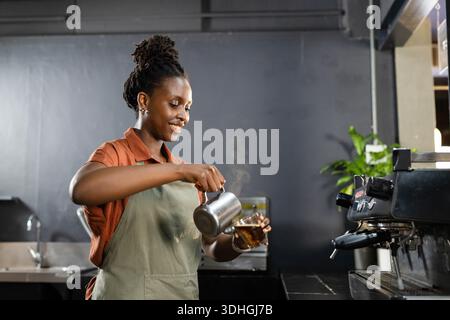 Afrikanische Barista trägt Olivenschürze und gießt aufgeschäumte Milch mit Milchkrug an der Kaffeekasse aus Stockfoto