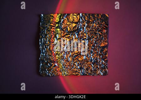 Zerknittertes Metallfolienquadrat auf abstraktem Neonverlaufshintergrund mit rotem Lichtbogen Stockfoto