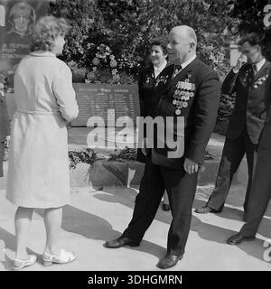 Slawjansk, Region Donezk, UdSSR. 1980er Jahre Ein Veteran aus dem Zweiten Weltkrieg mit einer Kiste voller Medaillen spricht mit einer Frau am AIZ-Werksdenkmal. Der ältere Mann lächelt, während er Erinnerungen teilt, während ein anderer Veteran im Hintergrund in der Nähe einer Steinplatte eine Träne aus dem Auge wischt, mit dem russischen Text „Ewige Herrlichkeit an dich“. Hinter ihnen steht das Denkmal mit einem Soldatenporträt. Dieses archivierte Schwarzweiß-Foto fängt die berührende, menschliche Seite der Feierlichkeiten zum Siegtag und die tiefen Emotionen der Generation der Sieger im friedlichen Donbass ein. Stockfoto