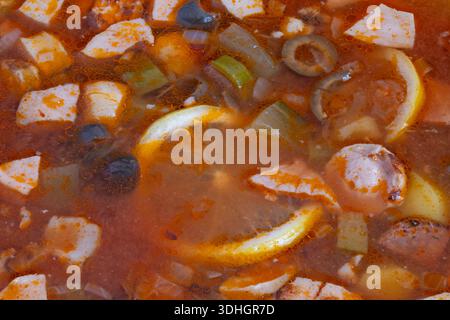Herzhafte Solyanka-Suppe mit Zitronenscheiben, Stückchen geräucherter Wurst, schwarzen Oliven, gewürfeltem Paprika und reichhaltiger Tomatenbrühe, in rustikaler Schüssel gemütlich zum Dämpfen Stockfoto
