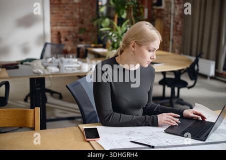Junge Erwachsene kaukasische Frau, die mit architektonischen Blaupausen an Laptop arbeitet Stockfoto
