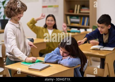 Kaukasisches Mädchen, das mit dem Kopf nach unten am Schreibtisch sitzt und von einer Gruppe von Klassenkameraden gemobbt wird, darunter kaukasischer Junge, asiatischer und kaukasischer Junge, der im Klassenzimmer zeigt und lacht Stockfoto