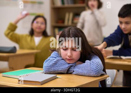Kaukasisches Mädchen, das am Schreibtisch sitzt, mit dem Kopf auf den Armen, das traurig erscheint, während Klassenkameraden im Hintergrund interagieren und ein Junge im Klassenzimmer auf sie zugreift Stockfoto