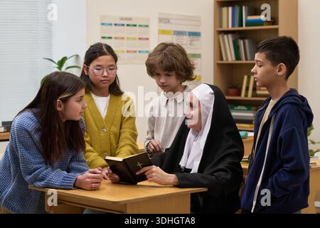 Kaukasische Nonne, die am Schreibtisch sitzt und Bibel einer Gruppe verschiedener Kinder und Teenager zeigt, darunter asiatische Mädchen und kaukasische Jungen, die alle aufmerksam im Klassenzimmer zuhören Stockfoto