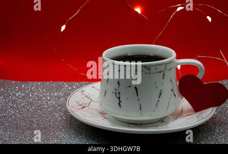 Kaffeetasse mit rotem Herz und warmen Feenlichtern auf rotem Glitzerhintergrund. Gemütliche Valentinstag-Stimmung für romantische Angebote. Stockfoto