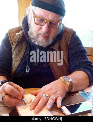 Senior-Mann mit Bartzeichnung in einem Notizbuch. Er trägt eine schwarze Mütze, eine braune Brille und ein mehrlagiges Outfit mit einer braunen Weste und einer Te Stockfoto