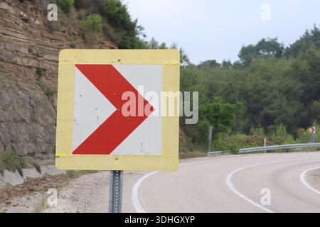 Nahaufnahme eines Verkehrswarnschildes mit dem roten Chevron Pfeil auf einer gewundenen Mountain Road mit einem felsigen Kliff und grünem Wald Hintergrund Stockfoto