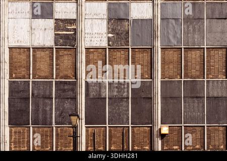 Sich wiederholendes Gitter aus verkleideten Industriefenstern mit nicht übereinstimmenden Holz- und Metallplatten, die Verfall, Verlassenheit und Widerstandsfähigkeit zeigen. Die Fassadenempha Stockfoto