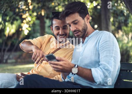 Zwei männliche Freunde, die zusammen auf ein Smartphone schauen, während sie im Park sitzen. Konzept der gemeinsamen digitalen Erfahrung, Technologie, Verbindung und Alltag Stockfoto