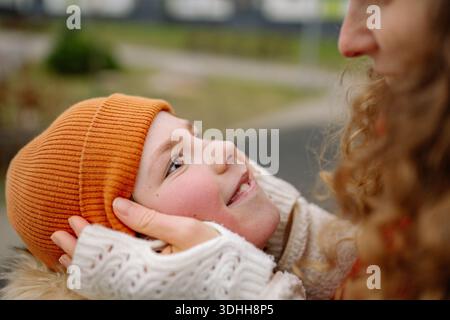Eine Frau und ein Kind lächeln einander an und zeigen eine enge Bindung. Sie stehen draußen in einem Park, tragen warme Hüte und gemütliche Kleidung. Die Szene erfasst einen Stockfoto
