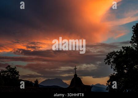 Dramatischer Sonnenuntergangshimmel mit glühenden Wolken über einem Kirchenkreuz und Bergsilhouetten. Eindrucksvolle mediterrane Abendszene in Corte, Korsika, Frankreich, Stockfoto