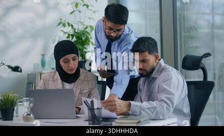 Business Arabian Team drei indische muslimische Kollegen im Büro diskutieren über Arbeitsprojekte mit Dokumenten, die Laptops mithilfe von digitalen Tablets betrachten Stockfoto