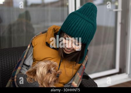 Eine junge Frau, die in eine Decke gewickelt ist und einen Winterhut trägt, hält und kuschelt draußen einen Yorkshire Terrier-Hund. Sie lächelt, während sie ihren Hund ansieht. Stockfoto