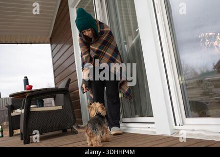 Eine lächelnde Frau mit grünem Hut und kariertem Schal spielt mit einem kleinen Yorkshire Terrier Hund auf einer hölzernen Veranda vor einem Haus. Das Wetter sieht bewölkt aus. Stockfoto