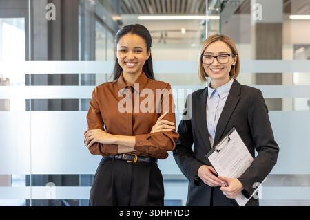 Zwei professionelle Geschäftsfrauen, eine mit überkreuzten Armen und eine andere mit einem Klemmbrett, stehen und lächeln selbstbewusst in einem modernen Glasbüro und zeigen Teamarbeit und Partnerschaft Stockfoto