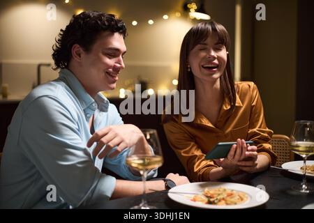 Kaukasischer junger Mann und kaukasische junge Frau sitzen am Tisch lachend, während sie während des Abendessens zusammen auf das Smartphone schauen, Weingläser und Teller mit Essen im Vordergrund sichtbar Stockfoto