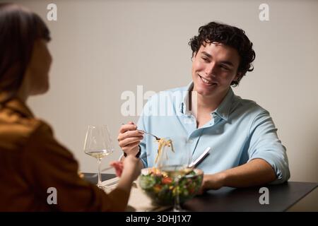 Junger kaukasischer Mann lächelt beim Nudelessen und sitzt der jungen Frau gegenüber am Tisch, hält die Gabel und sieht sie an, beide essen mit Weingläsern und Salatschalen Stockfoto