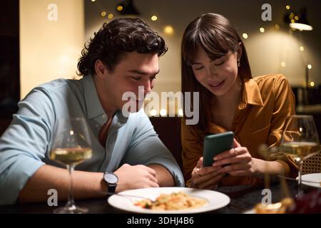 Junger kaukasischer Mann und junge kaukasische Frau sitzen zusammen am Tisch, lächeln und auf den Smartphone-Bildschirm schauen, essen mit Weingläsern und Essen im Vordergrund Stockfoto